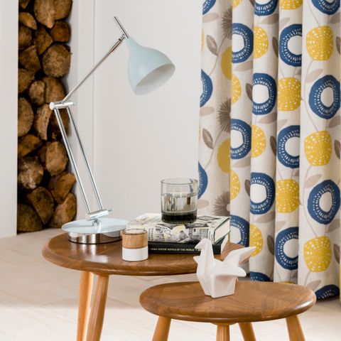 A set of tables with a white angle poise lamp with blue and yellow patterned curtains in the background