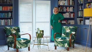 Erica Davies in front of white shutters in a blue living room with 2 arm chairs in green leaf patterned fabric