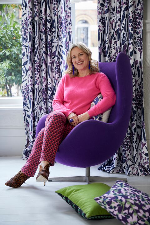 Sophie Robinson sat on a Purple Chair with matching colour curtains and cushions