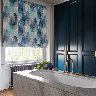 A blue bathroom with a large white bath in front of a window fitted with a Roman blind in Reflection Ocean fabric