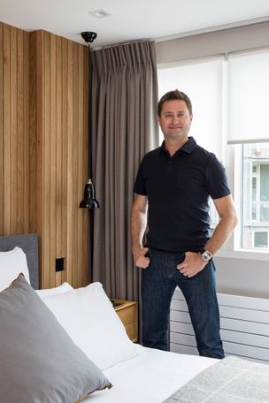 Acacia Ice motorised roller blinds fitted to rectangular windows and paired with bardot grey curtains in a bedroom with George Clarke standing front and centre