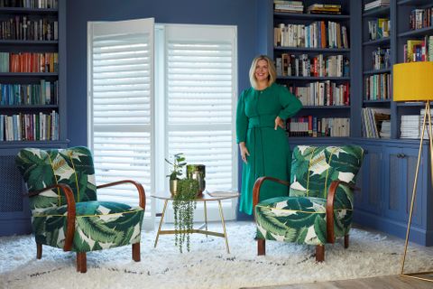 Erica Davies in front of white shutters in a blue living room with 2 arm chairs in green leaf patterned fabric