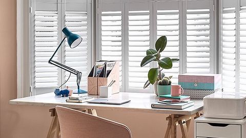White tier-on-tier Shutters in a bay window in a beige-themed office. There is a desk in front of the shutters.