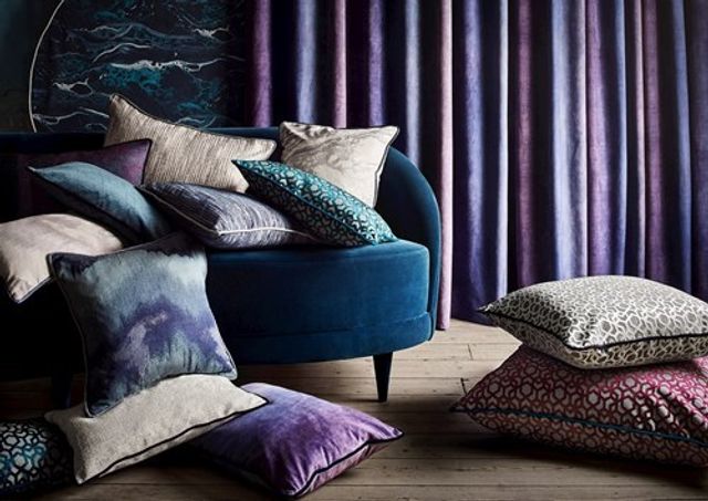 Cushions in varying shades of blue, purple and beige are piled on a blue arm chair with purple curtains in the background