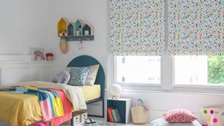 Children bedroom window dressed with roman blinds featuring multi-coloured dot pattern on a white background. Geometric printed cushions have been placed on blue bed and rug in the room.