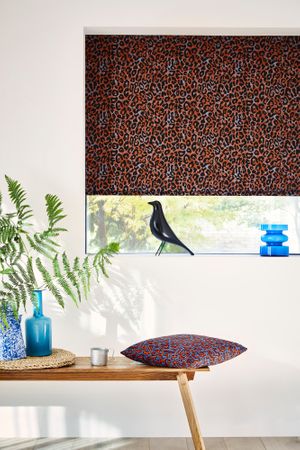 Boho Inspired black orange printed roman blinds are dressed on the windows in the room. Grey cushion featuring orange embroidery in retro pattern is placed on the table.