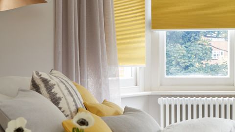 Corner close-up of bedroom where mustard yellow Thermashade blackout blinds at window, with Voile serene stone curtain hanging at the side. Matching mustard and stone cushions have been placed on the bed.
