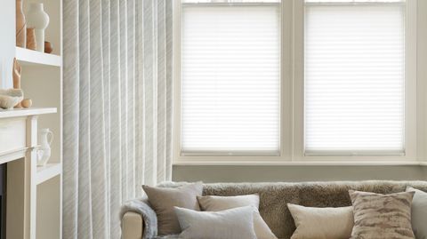Corner view of a living room where white curtains featuring light grey pattern on curtains dressed over cafe style Pleated blinds on windows.