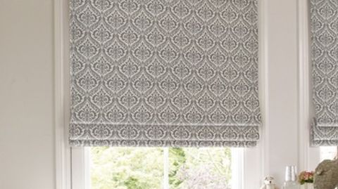 Silver and white roman blind designed with a repeating pattern is fitted to a rectangular window in a room decorated in white