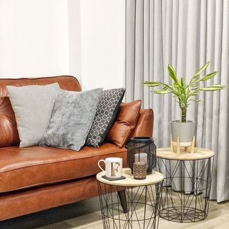 Light grey curtains against white coloured walls in a living room that has a brown leather sofa, small plant and a coffee table