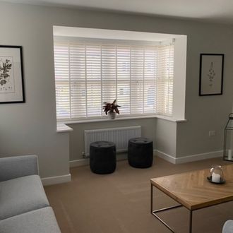 White shutters in a living room bay window with plant, two dark suede stools and a wooden coffee table