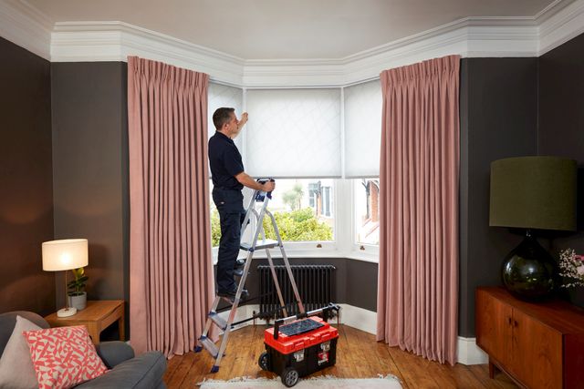 Advisor on ladder next to tool box installing white blinds behind pink curtains