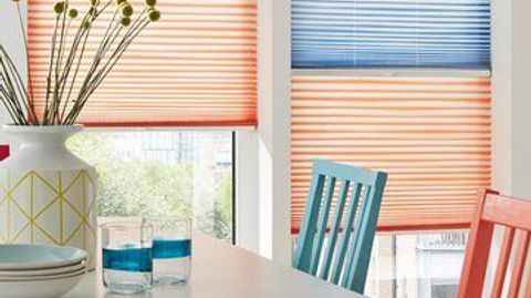 A tall white window has two pleated blinds fitted to the frame with the top blind in blue and the bottom blind in red they are in a dining room decorated in white that has two dining chairs in red and blue