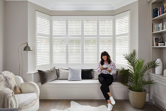 Anna Richardson sat in a white decorated living room with a bay window that has tiered silk white shutters