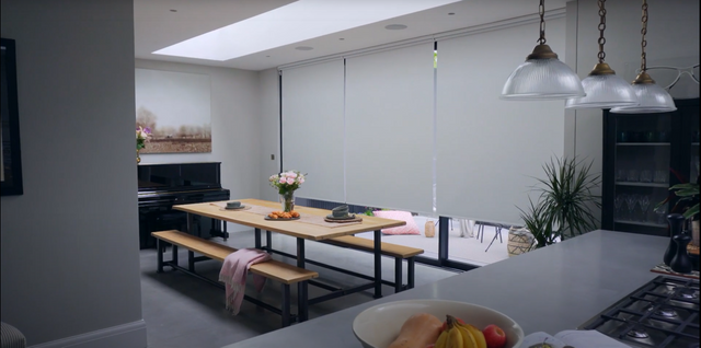 Electric roller blinds in a kitchen. There is a big dining table with chairs surrounding the table. There is a breakfast bar with a bowl of fruit on the table.