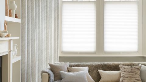 Corner view of a living room where white curtains featuring light grey pattern on curtains dressed over cafe style Pleated blinds on windows.