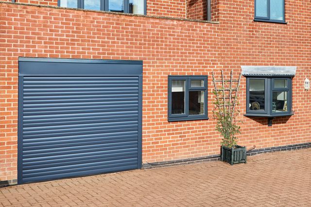 Angled shot of large red brick modern house with integral garage, with anthracite grey door, matching window frames and pipes, matching front door,, dark wooden planter with climber and brick driveway across the whole front of the house.