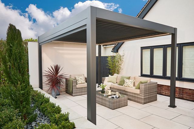 Exterior close up in a garden of a freestanding pergola in grey installed on a cream patio next to a cream rendered house and a raised flower border with alpine plants and a raffia style sofa and coffee table under the pergola.