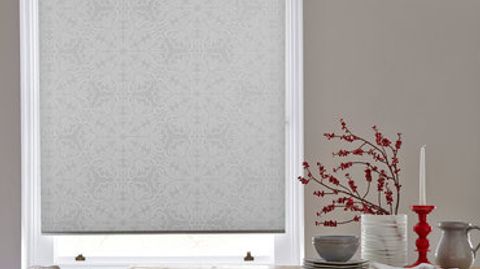 Silver coloured roller blind with a repeating pattern in a slightly darker shade attached to a tall rectangular window in a dining room that is decorated with beige walls and red blossom on the kitchen table