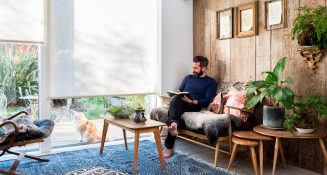 Oliver Heath reading a book in a living room that has floor to ceiling white roller blinds that are fitted to a door window