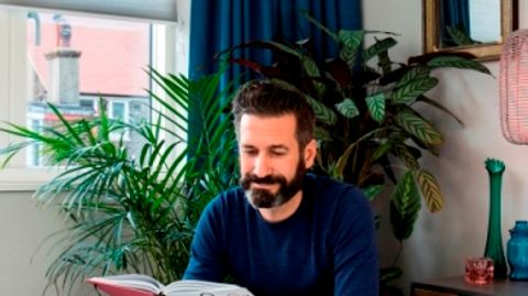 Oliver Heath sat reading a book in a room with a blue curtain and matching white roller blind