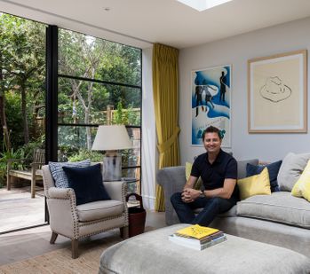 George Clarke sat on a couch in front of floor to ceiling Crittall windows with yellow curtains