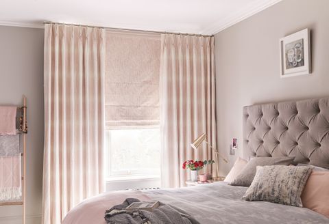 A bedroom featuring a bed in front of a window covered by a pink Roman blind under pink curtains