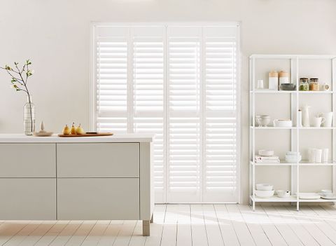 Tier on tier white shutters fitted to a rectangular shaped window in a kitchen with grey and white coloured kitchen cabinets, shelves, walls and wood flooring