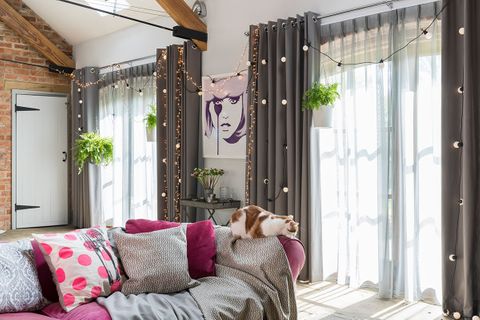 Grey eyelet curtains fitted to two rectangular windows in a living room with a purple sofa which has scatter cushions and a cat sitting on it