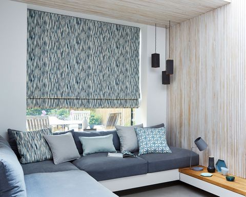 Roman curtains with a repeating pattern of light blue, dark blue and white fitted to a large rectangular window in a living room with a blue L shaped sofa and cushions and white decorated walls
