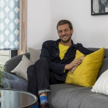 Charlie Luxton sat on a gray couch with yellow cushions 
