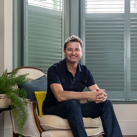George clarke sat on a cream chair in a living room that has teal coloured shutters