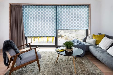 White and blue geometrically patterned roller blind matched with brown curtains on a wide door window in a living room with a grey sofa and armchair