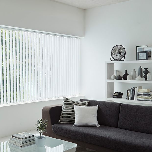 A close up of white vertical blinds in a white room with a grey sofa