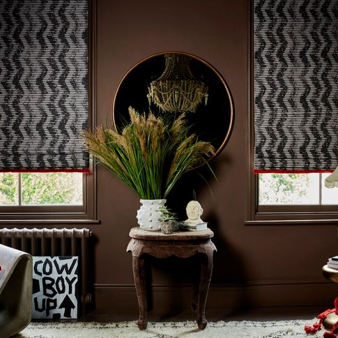 roman blinds with a black and white zigzag pattern and red fringing are fitted to tall windows in a room decorated with brown walls and plants on a antique table in the centre