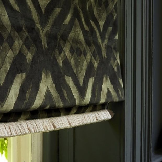 A dark green and black Roman blind finished in the Abigail Ahern Harkness Gasoline fabric. The blind has Colette Celeste fringing at the bottom