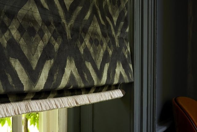 A dark green and black Roman blind finished in the Abigail Ahern Harkness Gasoline fabric. The blind has Colette Celeste fringing at the bottom