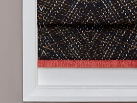 roman blind in black with white and gold patterns across the material along with red fringing is fitted to  a rectangular window 