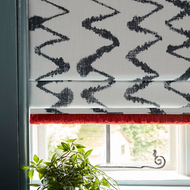 A white roman blind featuring a zig zag ink pattern down the full length of the blind along with red fringing and fitted on a blue framed window