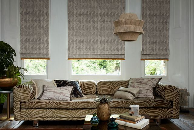 Beige patterned roman blinds with red fringing fitted to a series of windows in a living room with white windows and patterned sofa