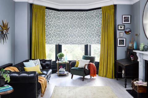 roman blinds with a repeating leaf and stem pattern in a teal colour, the blinds are matched with yellow curtains on a bay window in a living room decorated with grey walls and a black sofa  