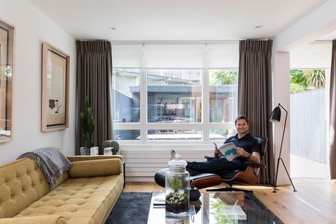 Interior Designer George Clarke sat in a black and brown armchair with a footrest in a living room featuring a yellow sofa, glass coffee table and grey curtains which are matched with white roller blinds that have been fitted to a wide rectangular window in the room
