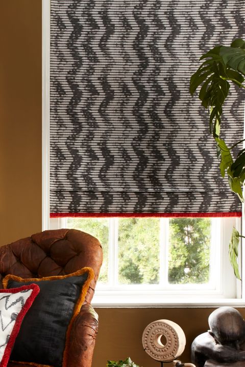 Black and white roman blinds with a zig zag pattern and red fringe is fitted to a rectangular window in a living decorated with brown walls