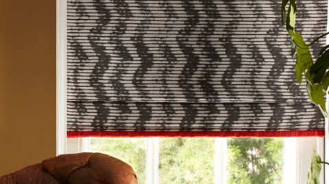Black and white roman blinds with a zig zag pattern and red fringe is fitted to a rectangular window in a living decorated with brown walls