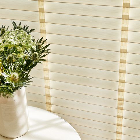 Close up of ivory wooden blinds with tape next to a side table with a vase of flowers 