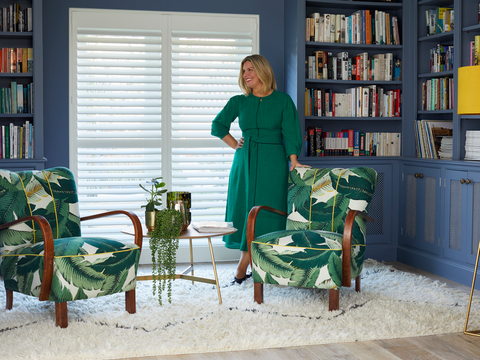 Erica Davies in front of white shutters in a blue living room with 2 arm chairs in green leaf patterned fabric