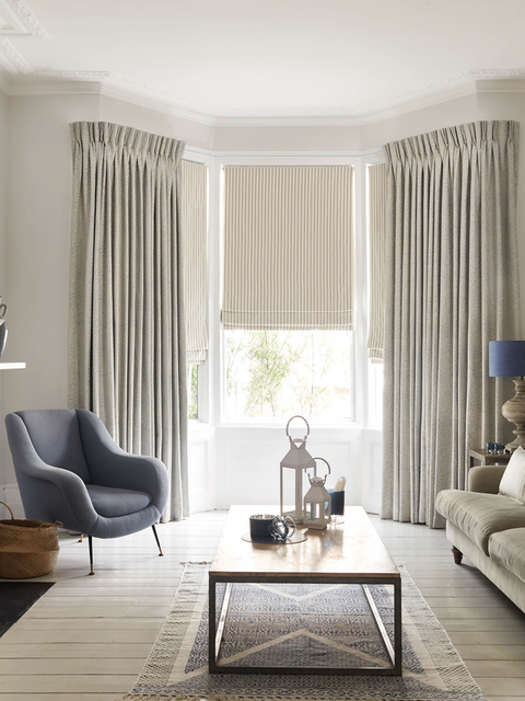 White curtains matched with cream roman blinds are fitted to bay shaped windows in a living room decorated in white
