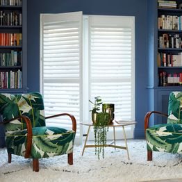full height silk white shutters on living room doors inbetween blue walls with bookcase behind green leaf themed armchairs and a table