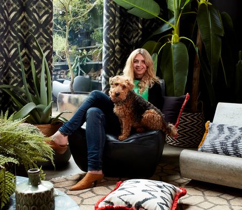Someone sat in a living decorated with plants and gasoline styled curtains from the abigail ahern collection while a dog sits in the centre  