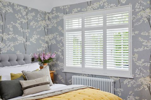 White tier on tier shutters fitted to a rectangular shaped winow in a bedroom decorated with grey and white floral wallpaper